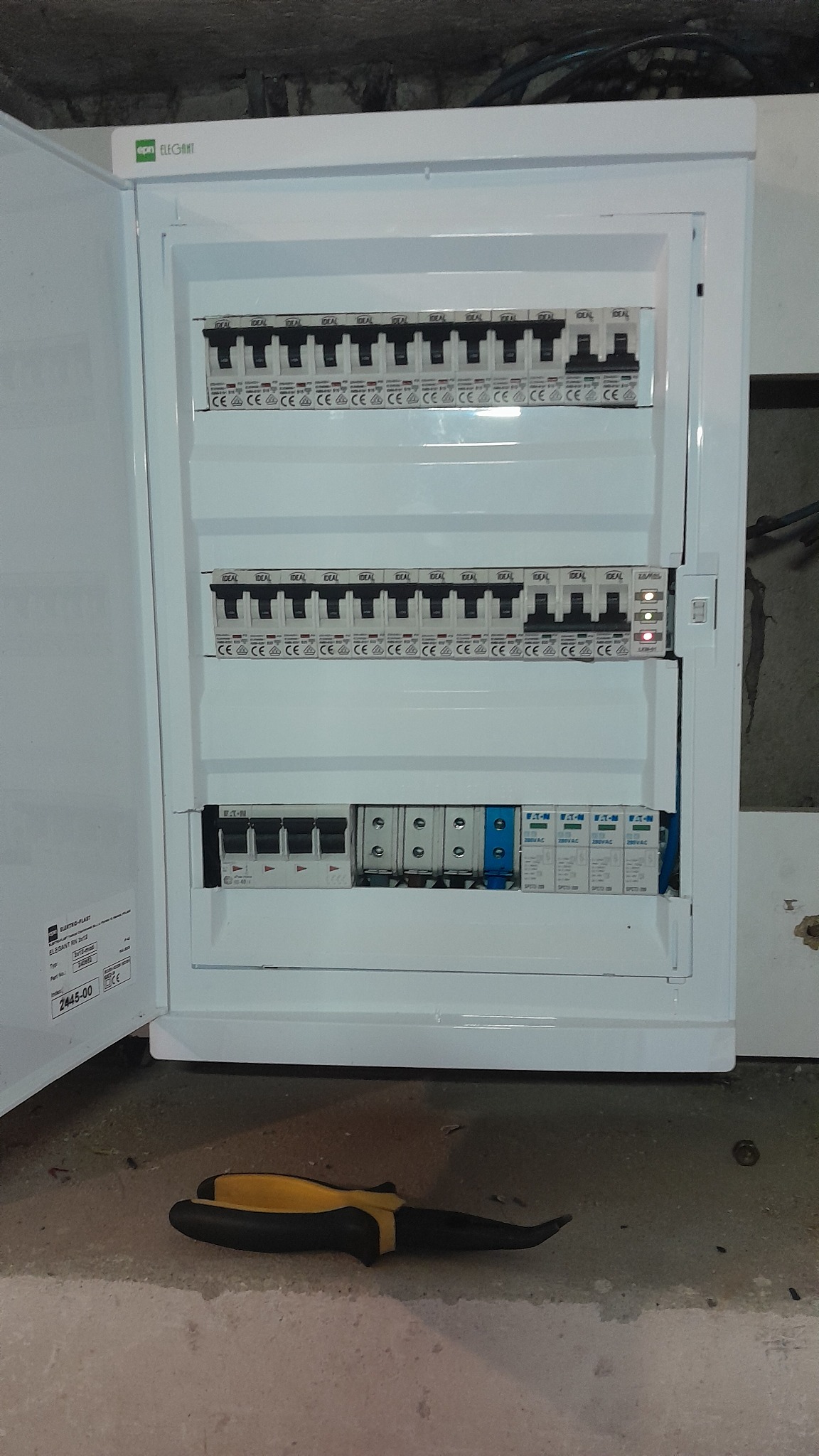 Otwarta biała skrzynka rozdzielcza z bezpiecznikami i wyłącznikami różnicowoprądowymi, widoczne oznaczenia producentów Ideal i Eaton, na podłodze leży czarno-żółty śrubokręt.