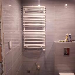 Osoba prywatna - Szare płytki w łazience z zamontowanym białym grzejnikiem drabinkowym, widoczna toaleta i niedokończone wykończenie ściany z lewej strony.