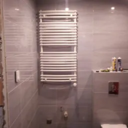 Szare płytki w łazience z zamontowanym białym grzejnikiem drabinkowym, widoczna toaleta i niedokończone wykończenie ściany z lewej strony.