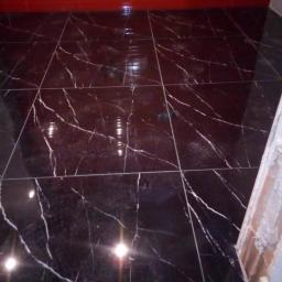 Osoba prywatna - Błyszcząca, czarna podłoga z płytek imitujących marmur z białymi żyłami, ułożona w łazience z czerwonymi płytkami na ścianie, widoczny fragment niedokończonej ściany z klejem do płytek.