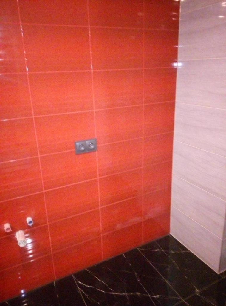 Pomarańczowe płytki ścienne z szarą fugą, szare gniazdko elektryczne, czarna marmurowa podłoga i fragment jasnoszarej ściany w rogu łazienki.