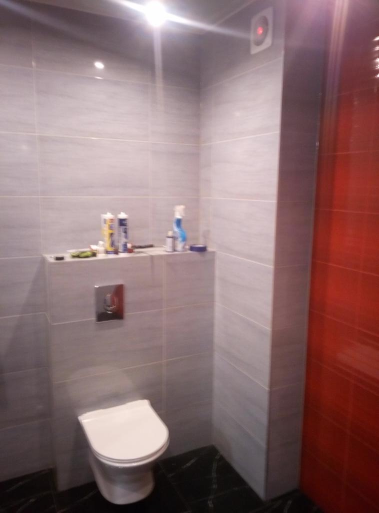 Wykończona łazienka z szarymi płytkami na ścianach i czarną podłogą, widoczna toaleta i akcesoria łazienkowe na półce.