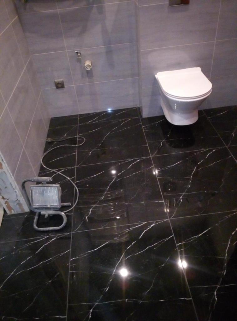 Wykończona łazienka z czarną marmurową podłogą i szarymi płytkami ściennymi, oświetlona lampą budowlaną, z widoczną toaletą podwieszaną.