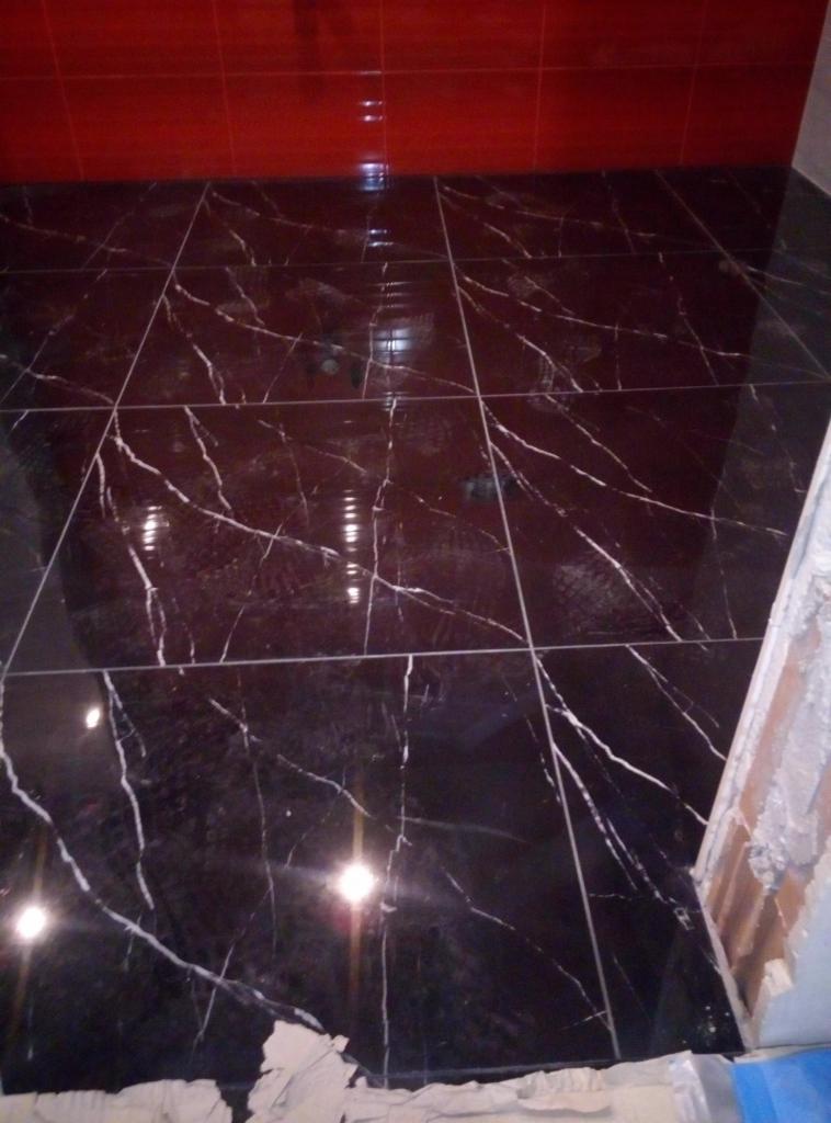 Błyszcząca, czarna podłoga z płytek imitujących marmur z białymi żyłami, ułożona w łazience z czerwonymi płytkami na ścianie, widoczny fragment niedokończonej ściany z klejem do płytek.