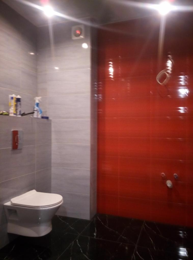 Nowoczesna łazienka z czerwonymi i szarymi płytkami, czarną marmurową podłogą i podwieszaną toaletą, widoczne elementy instalacji wodnej na czerwonej ścianie.