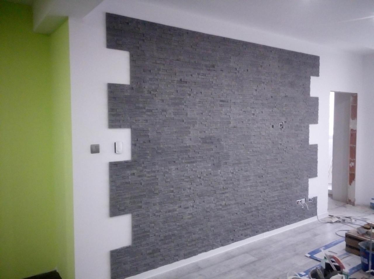 Ściana wyłożona szarą, dekoracyjną cegłą z nieregularnymi krawędziami, kontrastująca z białą ścianą i zielonym fragmentem ściany w rogu pomieszczenia. Widoczne gniazdka elektryczne i przełączniki.