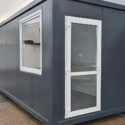 Szary kontener biurowy z białymi ramami okiennymi i drzwiami, widok z zewnątrz na kostce brukowej.