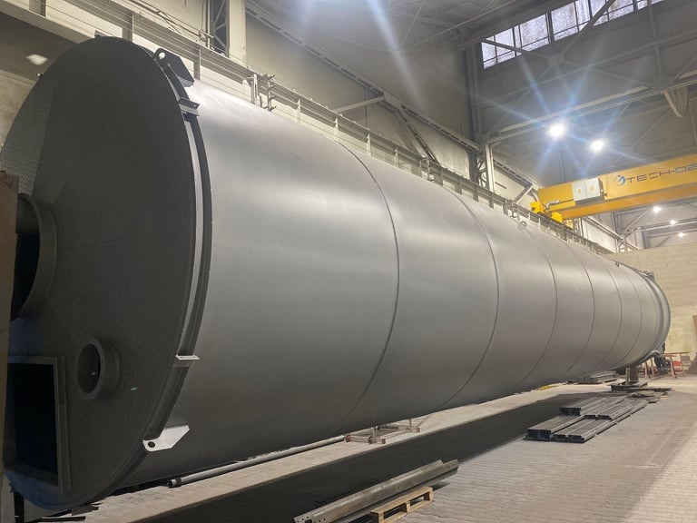 Duży, poziomy zbiornik przemysłowy o cylindrycznym kształcie, pomalowany na szaro, umieszczony w hali produkcyjnej z suwnicą w tle.