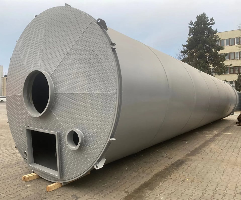 Duży, pomalowany na szaro, metalowy zbiornik cylindryczny z otworami o różnych kształtach, leżący na drewnianych belkach, na zewnątrz budynku.