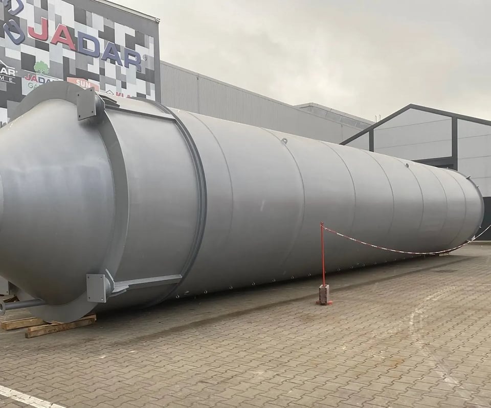 Duży, poziomy zbiornik przemysłowy w kolorze szarym, leżący na paletach na zewnątrz budynku z logo firmy Jadar.