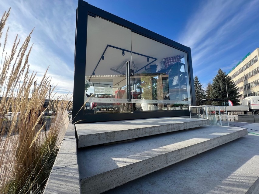 Nowoczesna, przeszklona konstrukcja z czarną ramą, umieszczona na betonowym podeście z kilkoma stopniami, widoczna z zewnątrz w słoneczny dzień, z elementami wyposażenia wnętrza widocznymi przez...