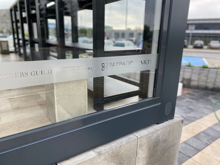 Szklana konstrukcja z czarnymi profilami, oklejona folią z logotypami firm, osadzona na betonowym fundamencie na zewnątrz budynku.