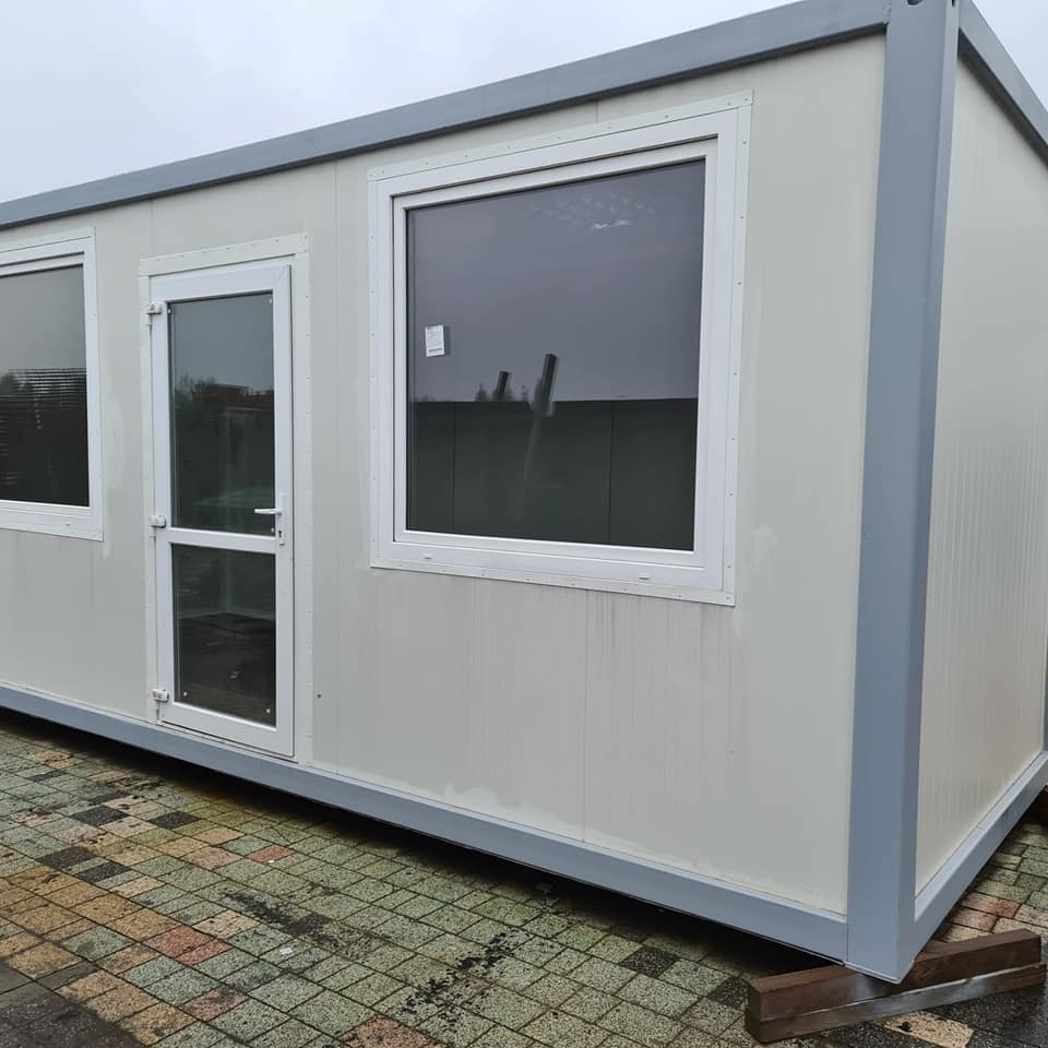 Gotowy kontener biurowy z oknami i drzwiami, ustawiony na drewnianych legarach na kostce brukowej w pochmurny dzień.