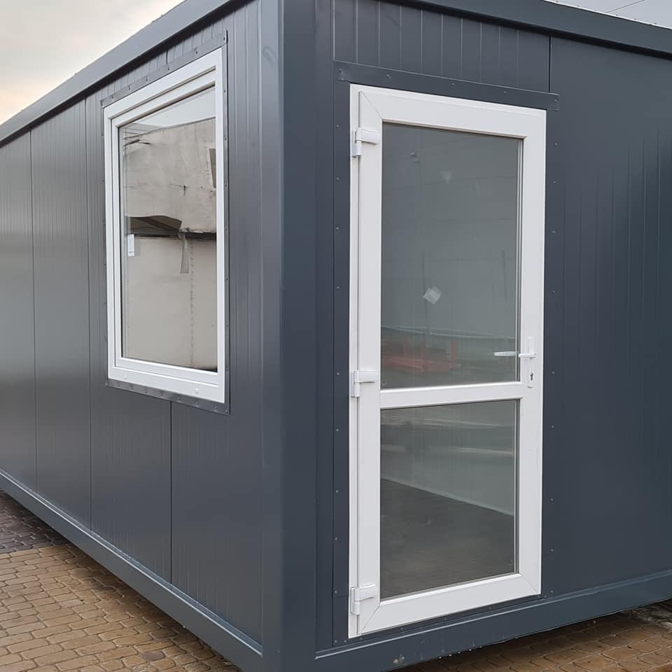 Szary kontener biurowy z białymi ramami okiennymi i drzwiami, widok z zewnątrz na kostce brukowej.