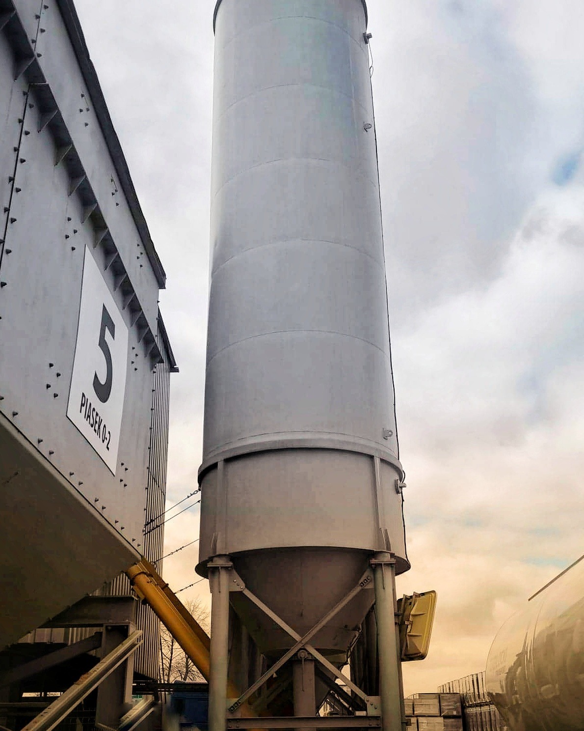 Dwa metalowe silosy na piasek o różnej konstrukcji, jeden z numerem 5 i napisem 'Piasek 0-2', drugi cylindryczny, na tle pochmurnego nieba.