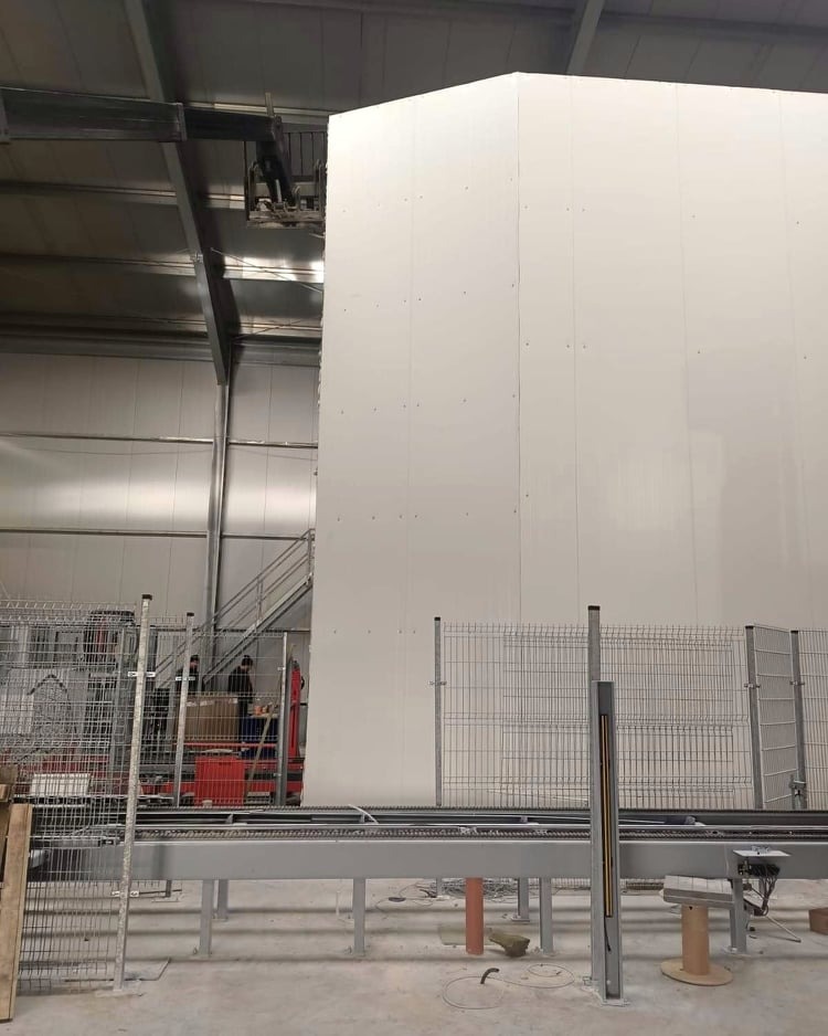 Wnętrze hali produkcyjnej z widoczną konstrukcją z białych paneli, metalowe ogrodzenia oraz elementy linii produkcyjnej z podajnikiem taśmowym.