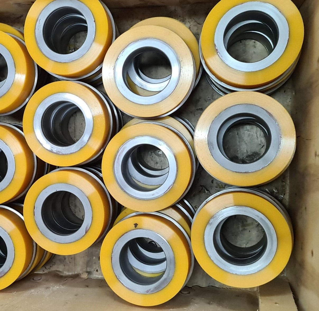 Stos żółto-srebrnych pierścieni z metalu i tworzywa sztucznego, ułożonych w kartonowym pudełku, potencjalnie uszczelki lub elementy maszyn.