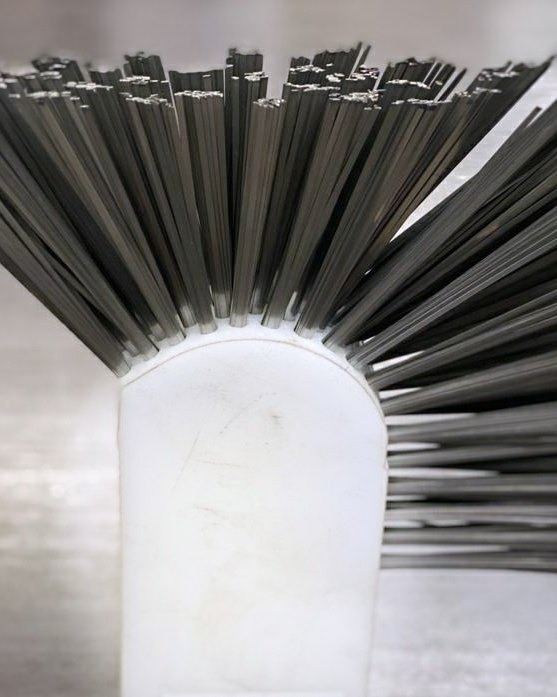 Szczotka druciana z białym uchwytem, widoczne ślady użytkowania na uchwycie, ustawiona pionowo, włosie skierowane ku górze.