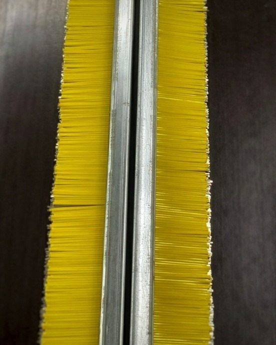 Szczotka przemysłowa z żółtym włosiem zamocowana w metalowej ramie z widocznym rowkiem pośrodku.