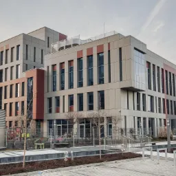 Nowoczesny budynek biurowy z elementami w kolorze ceglastym, widok z zewnątrz, z cylindrycznymi elementami wentylacyjnymi na pierwszym planie i logo firmy na dachu.