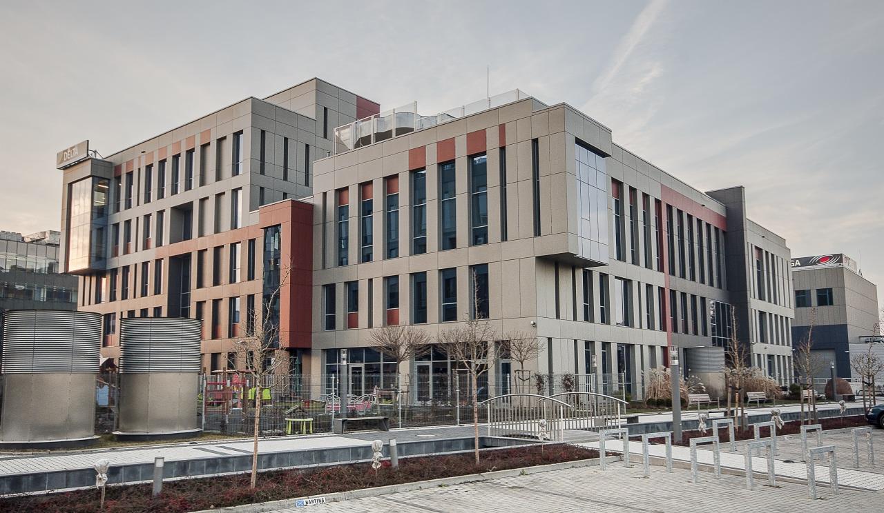 Nowoczesny budynek biurowy z elementami w kolorze ceglastym, widok z zewnątrz, z cylindrycznymi elementami wentylacyjnymi na pierwszym planie i logo firmy na dachu.