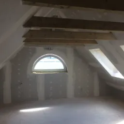 Poddasze w trakcie wykańczania z zamontowanymi płytami na ścianach i suficie, widoczne drewniane belki konstrukcyjne i okna dachowe, ślady po szpachlowaniu na płytach.