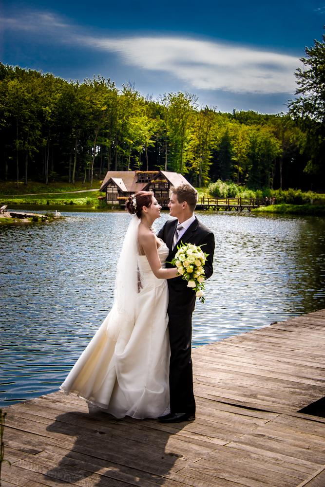 Para młoda na drewnianym pomoście nad jeziorem, w tle leśny krajobraz i domek z pomostem, panna młoda w białej sukni trzyma bukiet żółtych róż, oboje patrzą na siebie z uśmiechem.