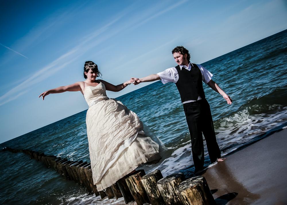 Para młoda trzymająca się za ręce na plaży, panna młoda w sukni ślubnej koloru ecru, pan młody w czarnej kamizelce, drewniane pale w wodzie wzdłuż brzegu, błękitne niebo w tle, lekki wiatr i fale.