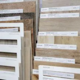 Ekspozycja próbek płytek podłogowych i ściennych w różnych odcieniach beżu i brązu, z widocznymi etykietami cenowymi i nazwami produktów, ułożonych pionowo na stojaku w sklepie.