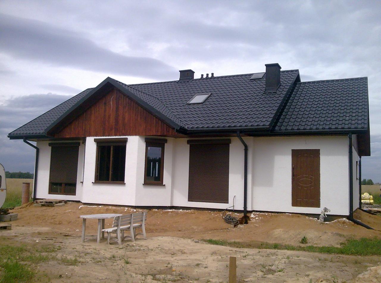 Parterowy dom szkieletowy z ciemną dachówką i drewnianą elewacją szczytową, widoczne rolety zewnętrzne i drewniane drzwi wejściowe, teren wokół domu w trakcie zagospodarowywania.