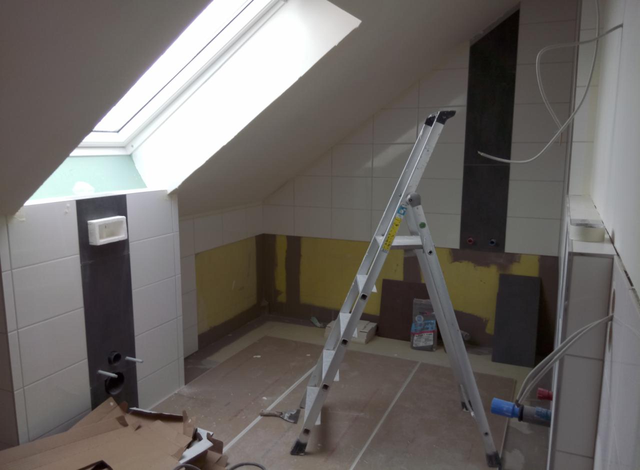 Łazienka na poddaszu w trakcie remontu, widoczna drabina, częściowo położone płytki, surowe ściany, okno dachowe w skosie.