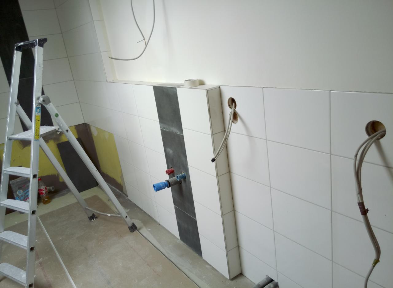 Łazienka w trakcie remontu: białe płytki, czarny pas dekoracyjny, wystające rury i kable, drabina aluminiowa, widoczne przygotowania do instalacji armatury.