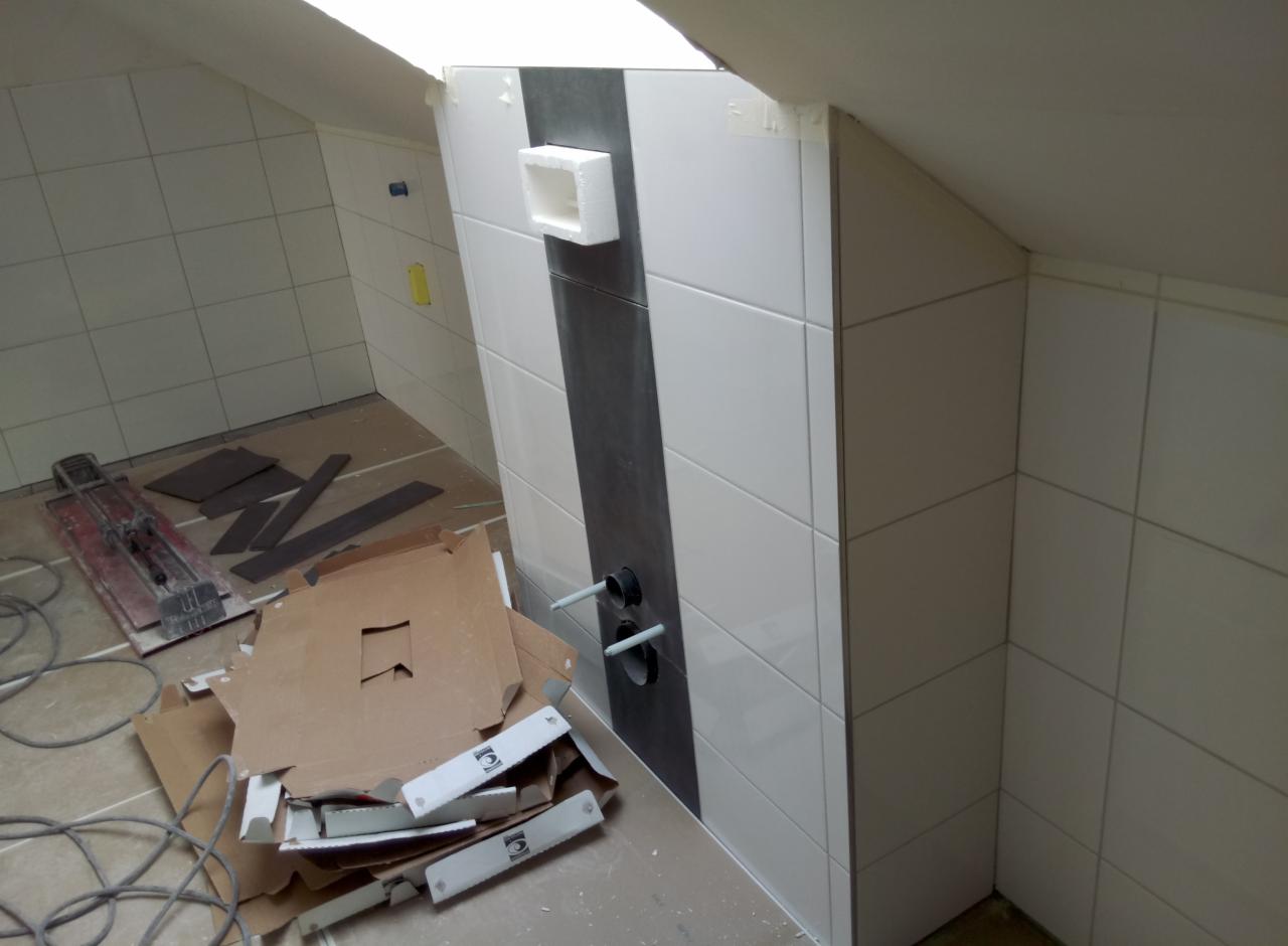 Łazienka na poddaszu w trakcie wykańczania: białe płytki z szarym pasem, wystające rury instalacji, kartony po płytkach, gilotyna do cięcia płytek.