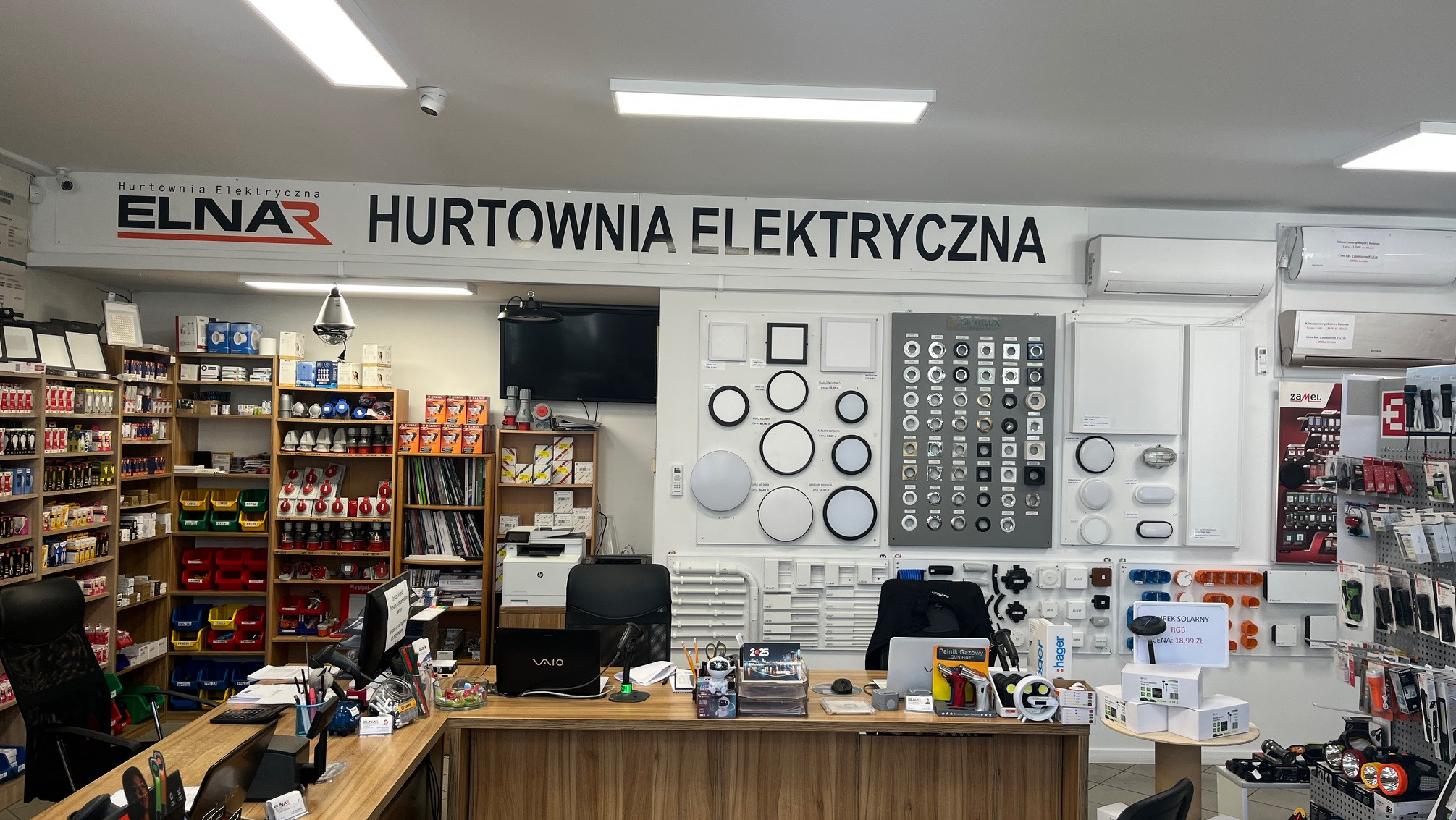 Wnętrze hurtowni elektrycznej Elnar: regały z asortymentem, ekspozycja lamp i akcesoriów oświetleniowych, biurko obsługi z laptopem Vaio.