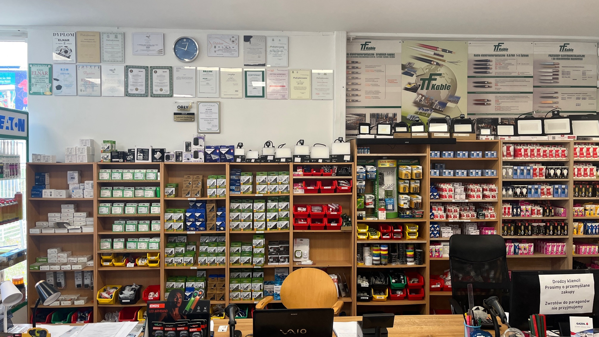 Wnętrze sklepu hydraulicznego: półki z osprzętem elektrycznym, oświetleniem i kablami. Na ścianie dyplomy i reklamy kabli TF. Widok zza lady.