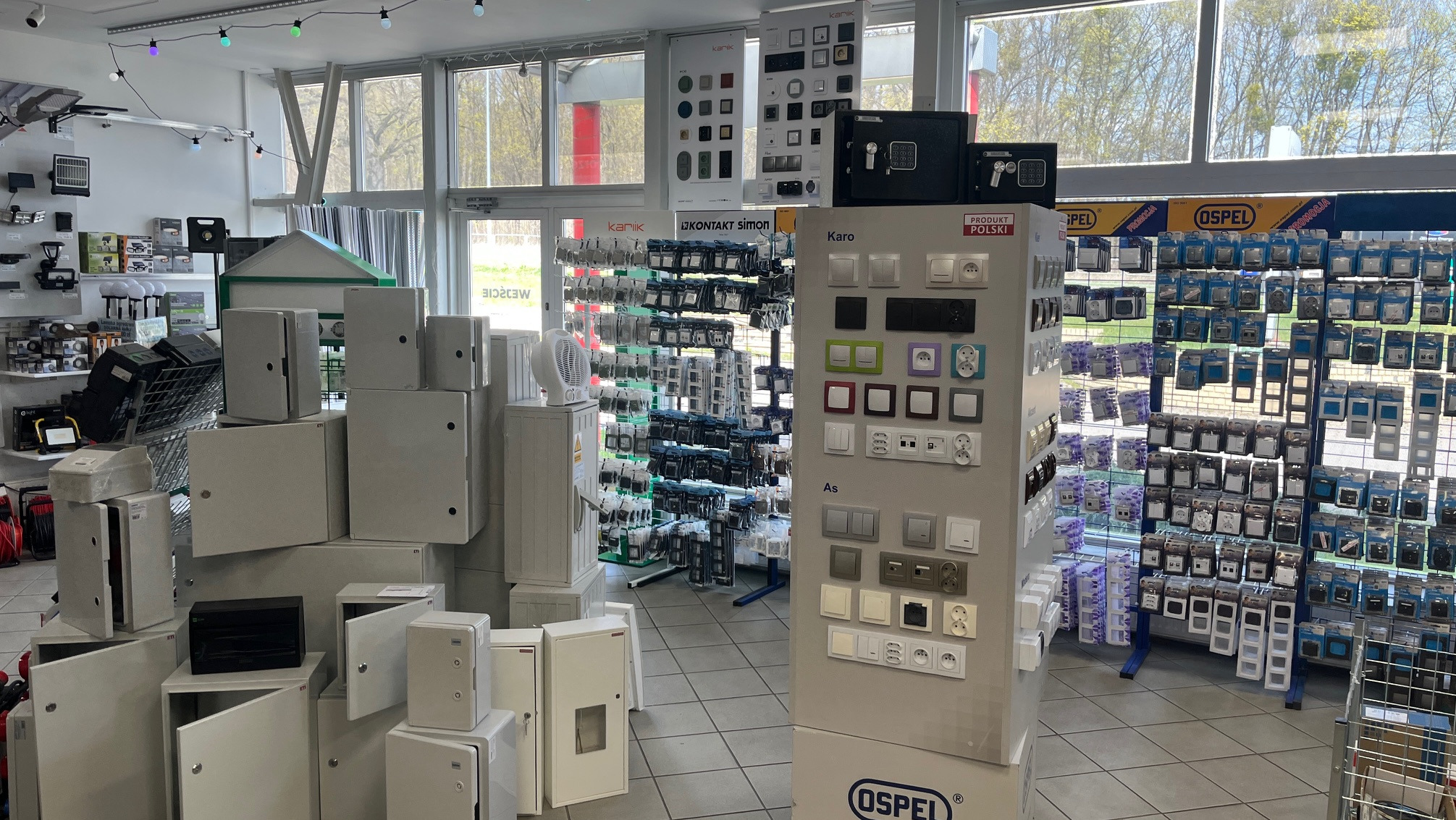Wnętrze sklepu hydraulicznego: ekspozycja rozdzielnic elektrycznych, włączników i gniazdek różnych marek (Kanik, Kontakt Simon, Ospel) oraz akcesoriów instalacyjnych.