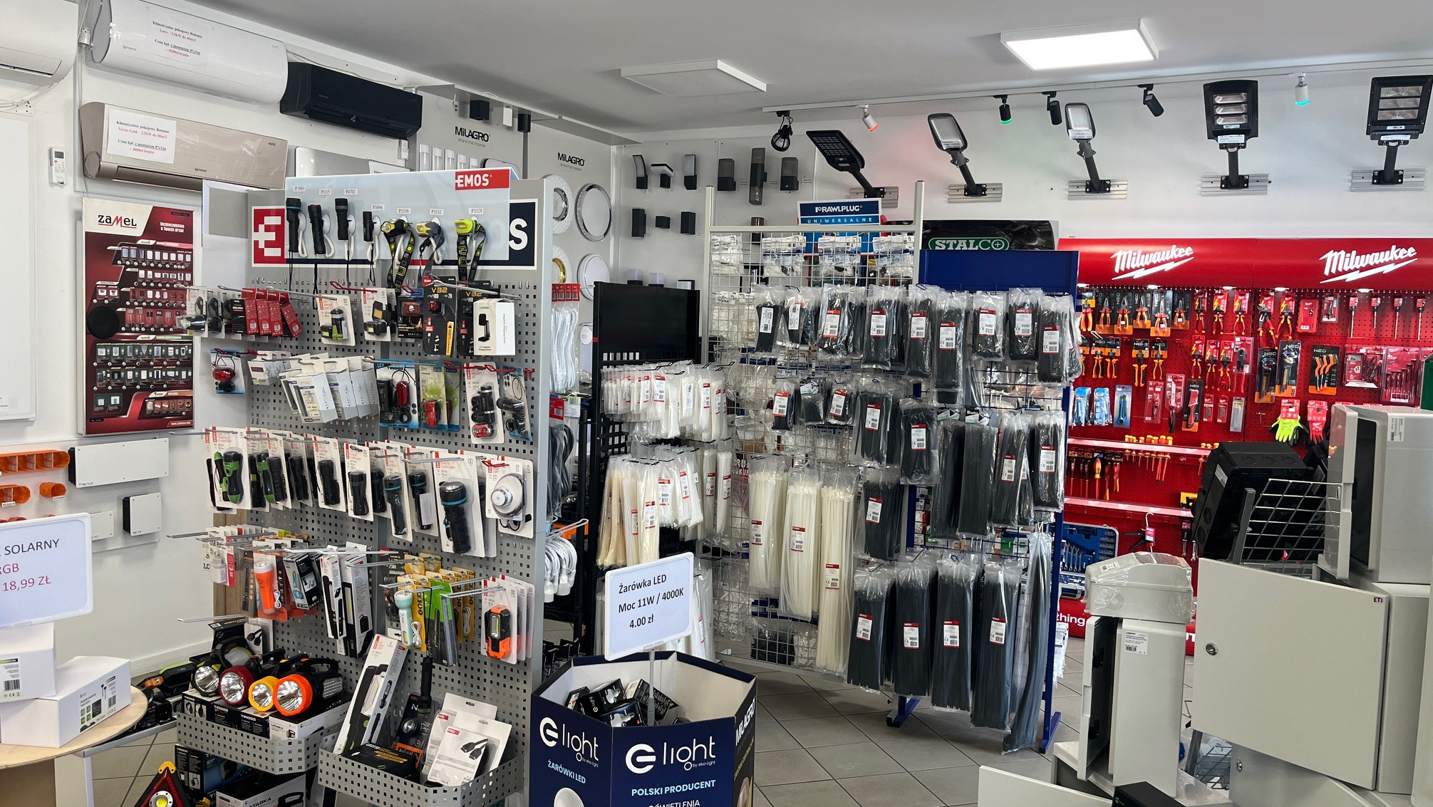 Ekspozycja sklepu z oświetleniem LED, narzędziami i akcesoriami instalacyjnymi. Widoczne lampy, latarki, oprawy oświetleniowe, narzędzia ręczne i elementy instalacyjne.