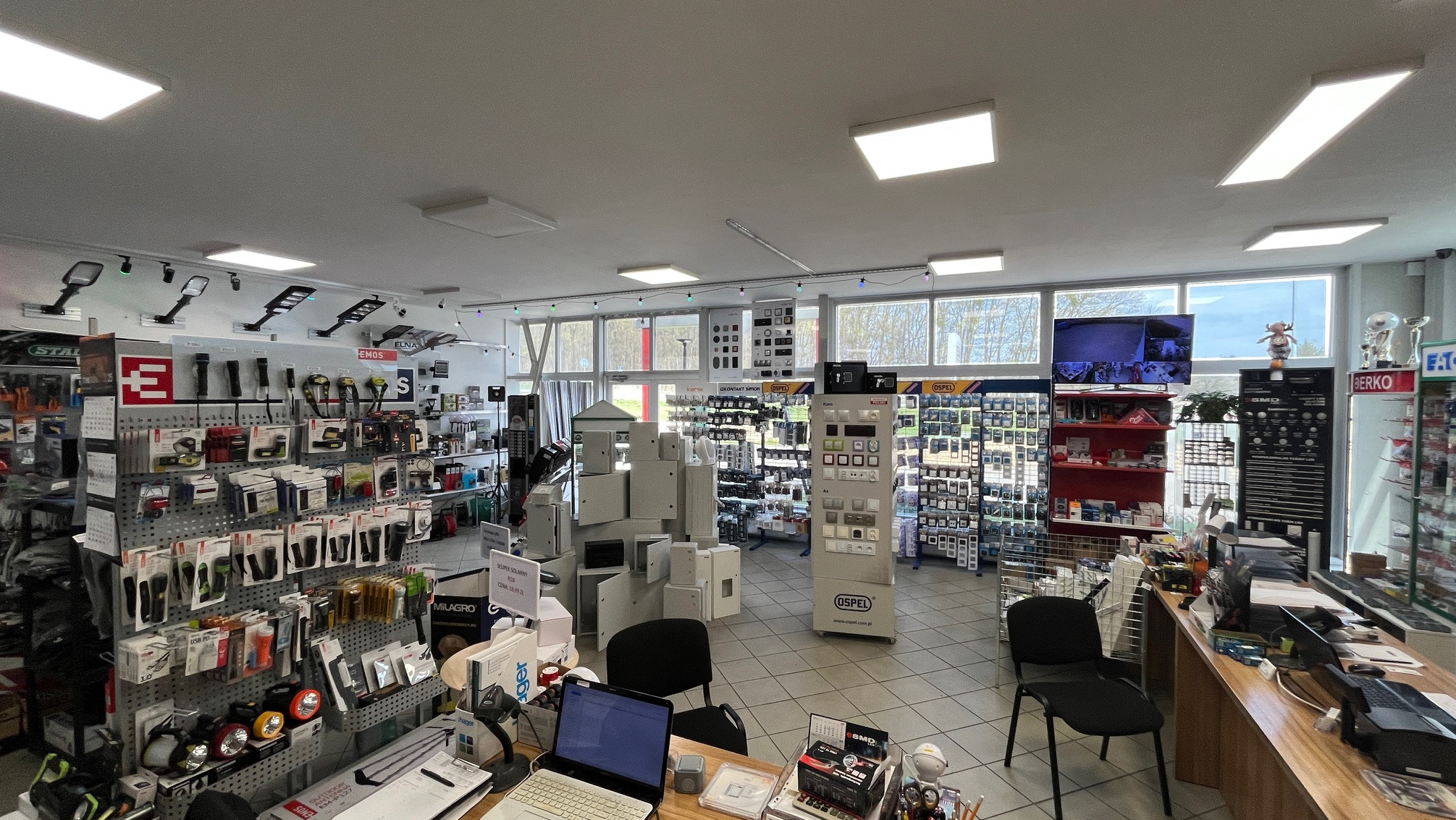 Wnętrze sklepu z artykułami elektrycznymi: ekspozycja oświetlenia LED, przełączników Ospel i akcesoriów. Widok zza lady, laptop na pierwszym planie. Jasne wnętrze, okna w tle.