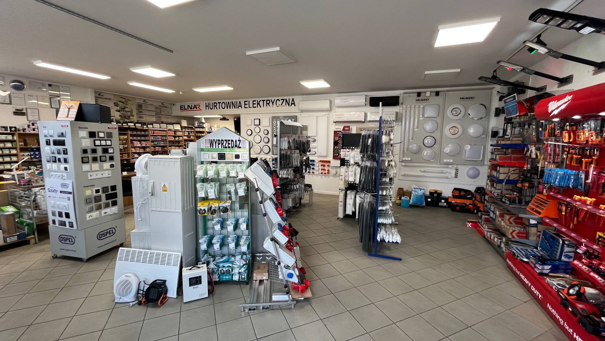 Wnętrze sklepu elektrycznego w Chełmie. Ekspozycja z włącznikami Ospel, oświetleniem Milagro i narzędziami Milwaukee. Widoczny napis ELNAR Hurtownia Elektryczna.