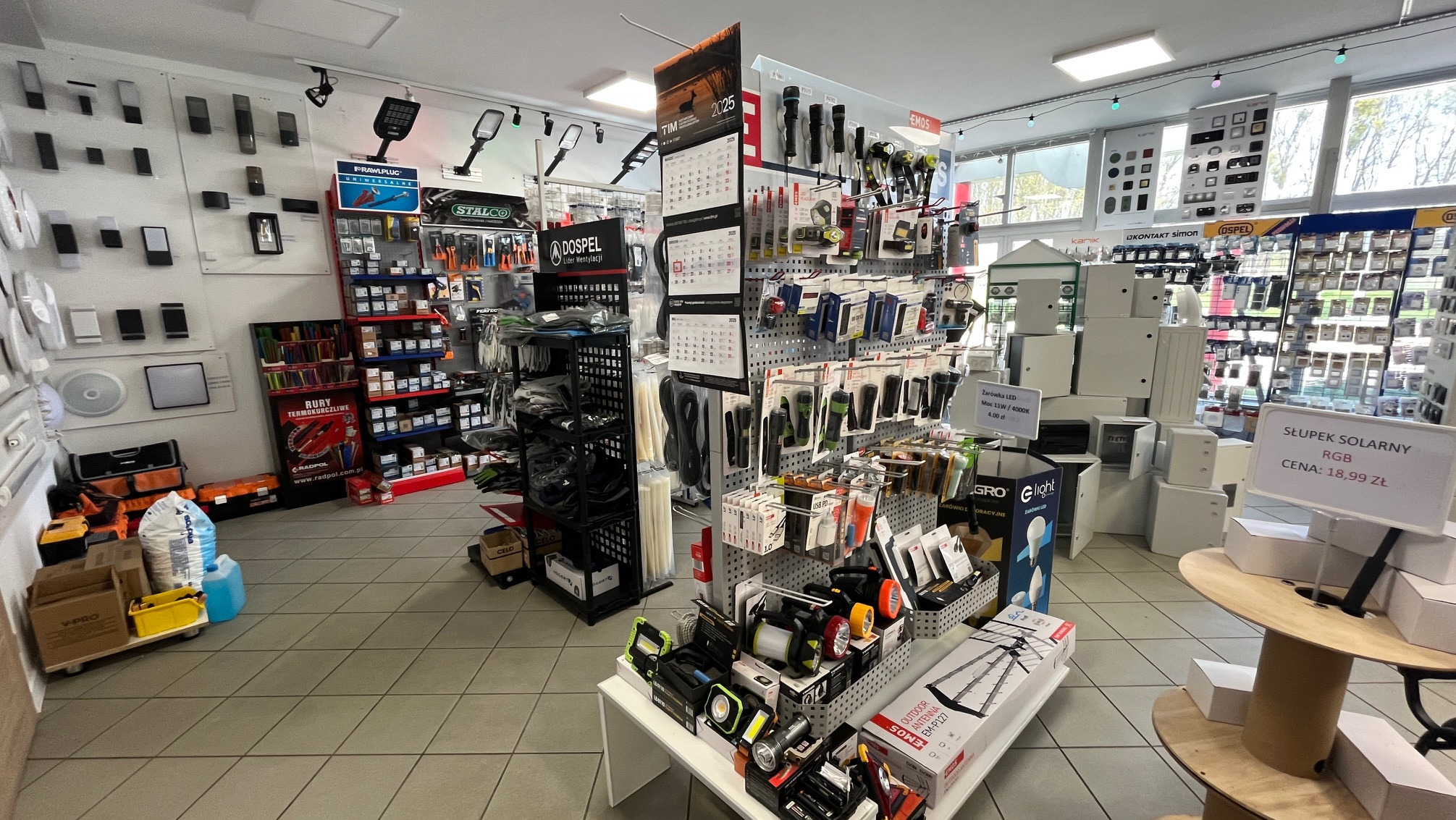 Wnętrze sklepu z oświetleniem i akcesoriami elektrycznymi. Widoczne półki i ekspozytory z różnorodnym asortymentem, w tym lampy solarne, żarówki LED i elementy instalacyjne.