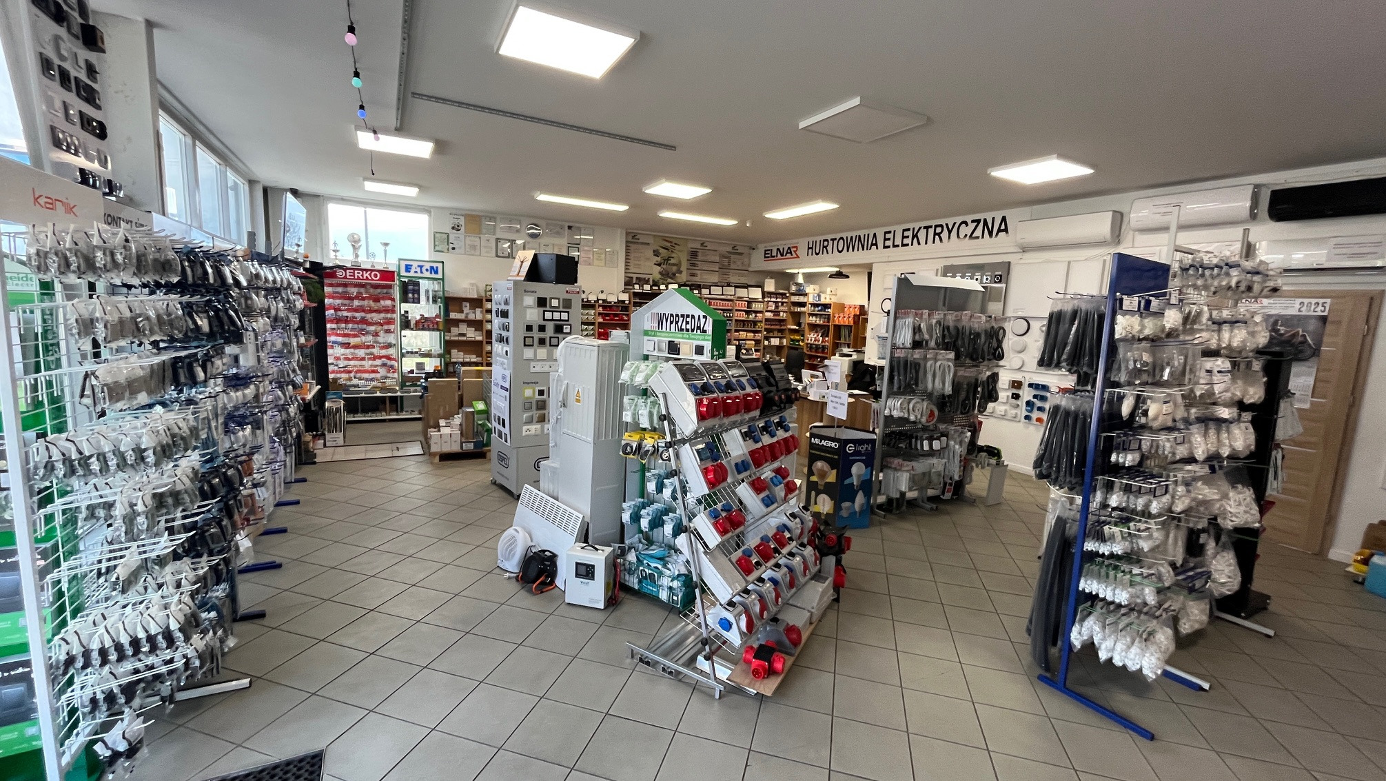 Wnętrze sklepu elektrycznego: regały z osprzętem, wyłączniki, kable, gniazda, oświetlenie. Widoczny napis 'Hurtownia Elektryczna ELNAR'.