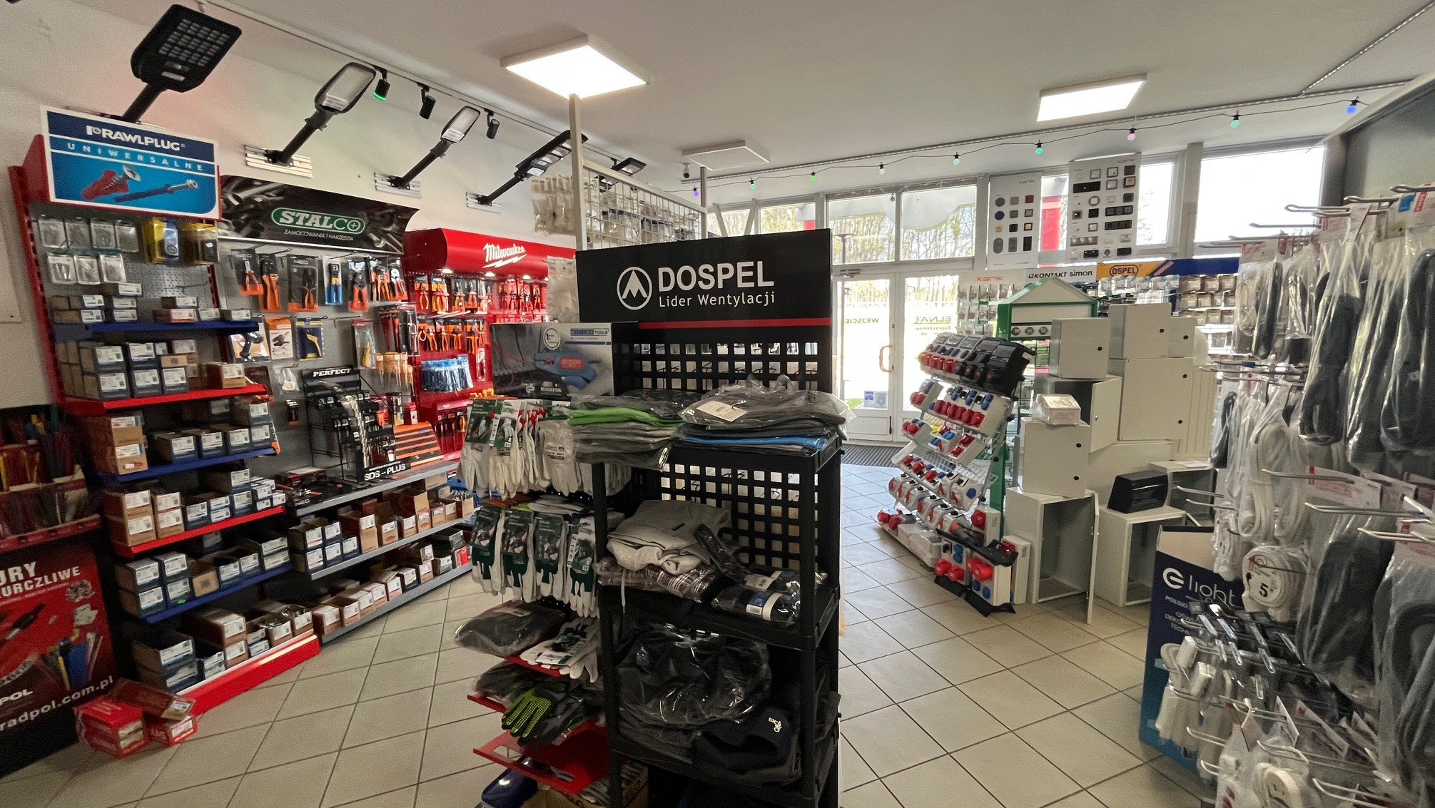 Wnętrze sklepu instalacyjnego: regały z narzędziami, materiałami montażowymi i akcesoriami wentylacyjnymi marki Dospel. Widok na ekspozycję z oświetleniem.