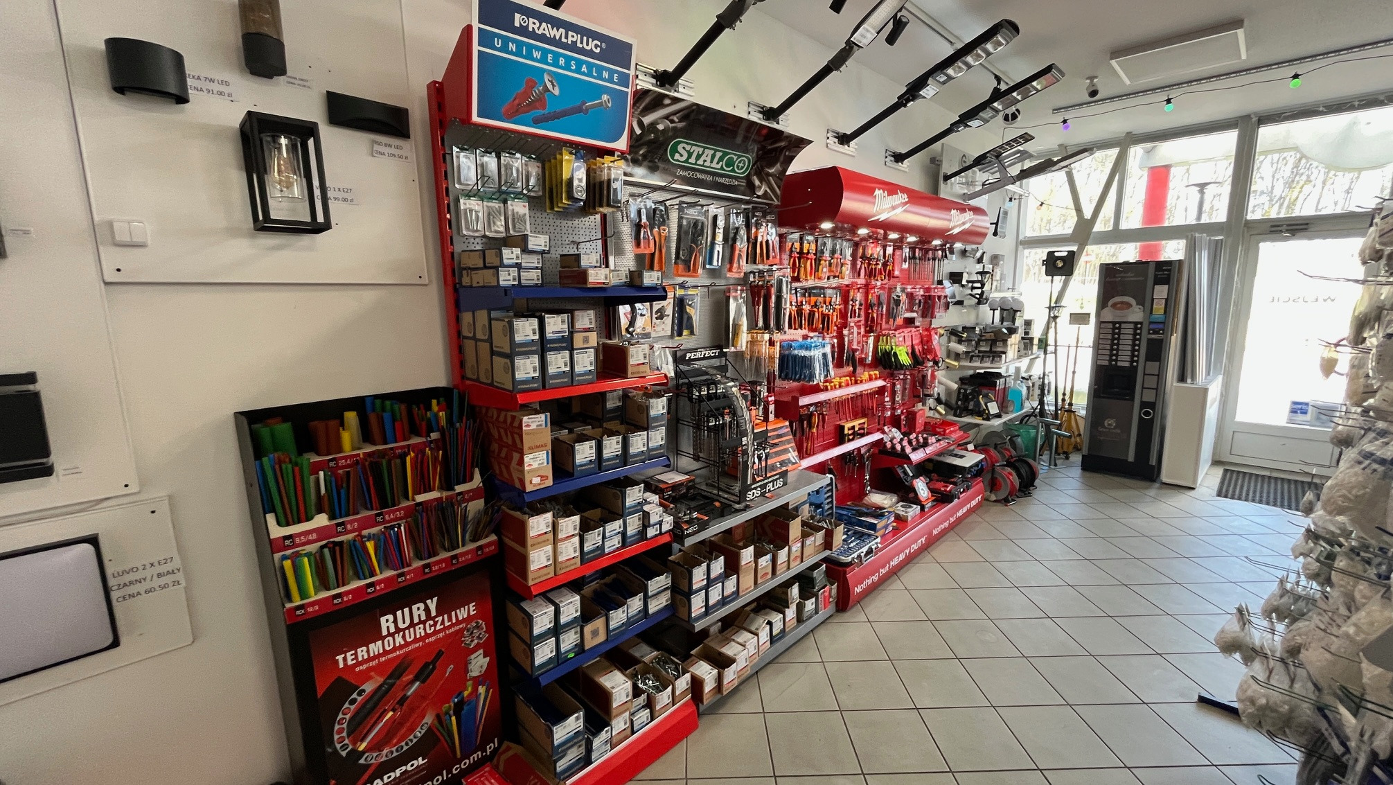 Ekspozycja w sklepie hydraulicznym: narzędzia Stalco, rury termokurczliwe Adpol, uniwersalne kołki Rawlplug. Oświetlenie i automat z kawą w tle.