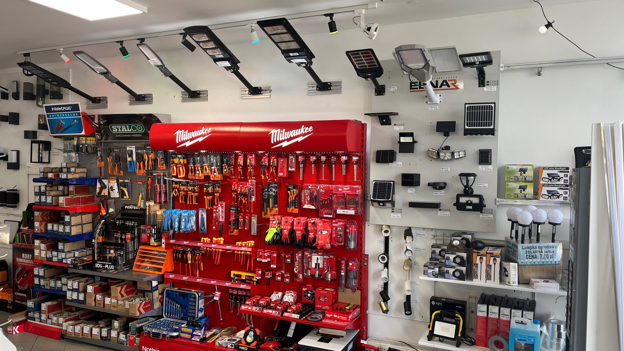 Ekspozycja w sklepie: narzędzia Milwaukee, oświetlenie LED, lampy solarne ogrodowe. Różnorodność produktów na półkach i ścianach, z czerwonym regałem Milwaukee w centrum.