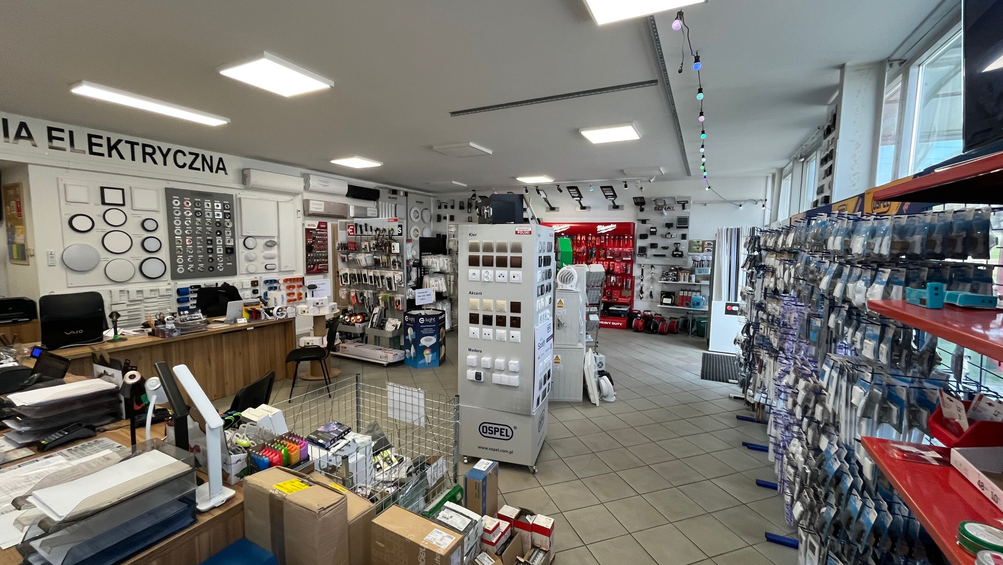 Wnętrze sklepu elektrycznego z ekspozycją gniazdek Ospel, oświetlenia i narzędzi. Widoczne regały z akcesoriami i stanowisko obsługi klienta. Jasne oświetlenie.