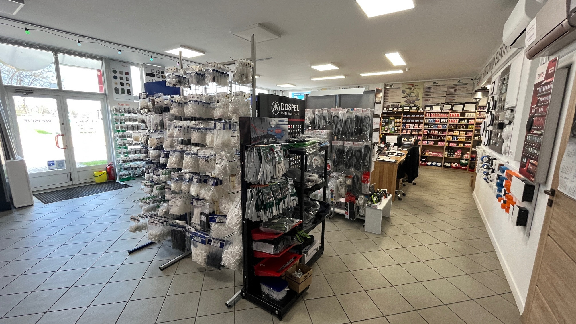 Wnętrze sklepu z asortymentem instalacyjnym: rury, złączki, narzędzia i akcesoria wentylacyjne na regałach, z logo Dospel. Jasne oświetlenie, czyste podłogi.