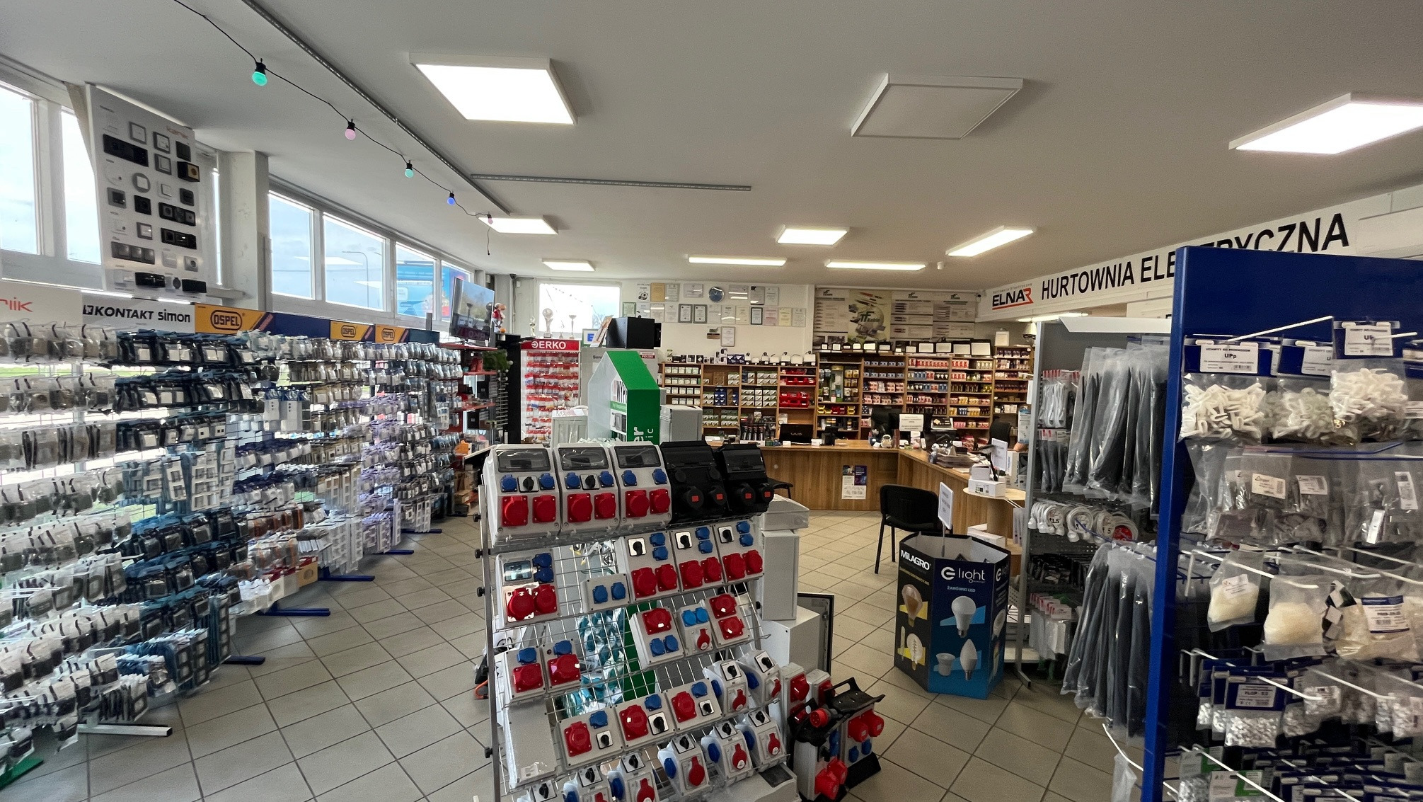 Wnętrze sklepu elektrycznego: regały pełne akcesoriów, gniazda, wtyczki, kable. Widoczny napis 'ELNAR Hurtownia Elektryczna'. Jasne oświetlenie, czyste wnętrze.