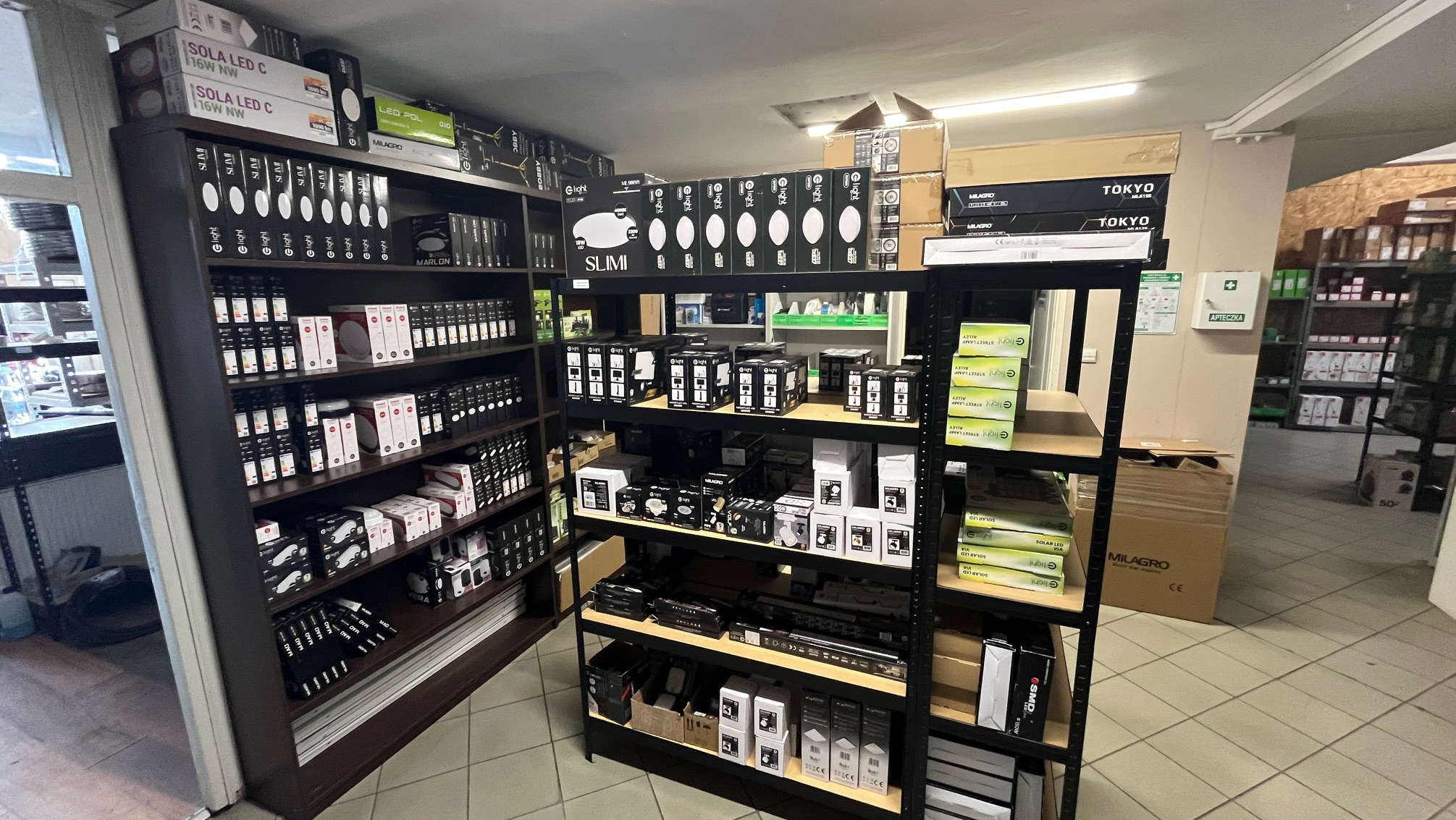 Oświetlenie LED na półkach sklepowych: lampy sufitowe, panele, reflektory. Różne modele i marki, np. Milagro, E-light, Tokyo. Apteczka na ścianie w tle.