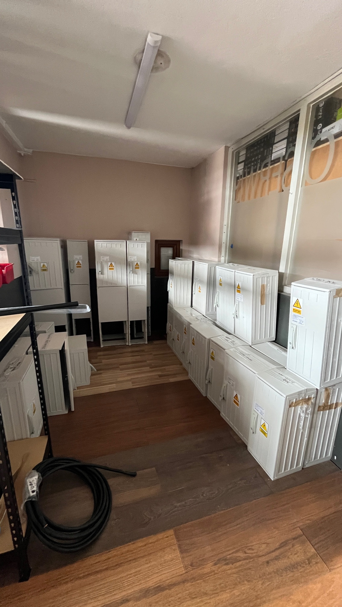 Magazyn białych skrzynek z symbolem ostrzegawczym, ułożonych rzędami na podłodze z paneli. W tle widoczne dodatkowe skrzynki i fragment regału.