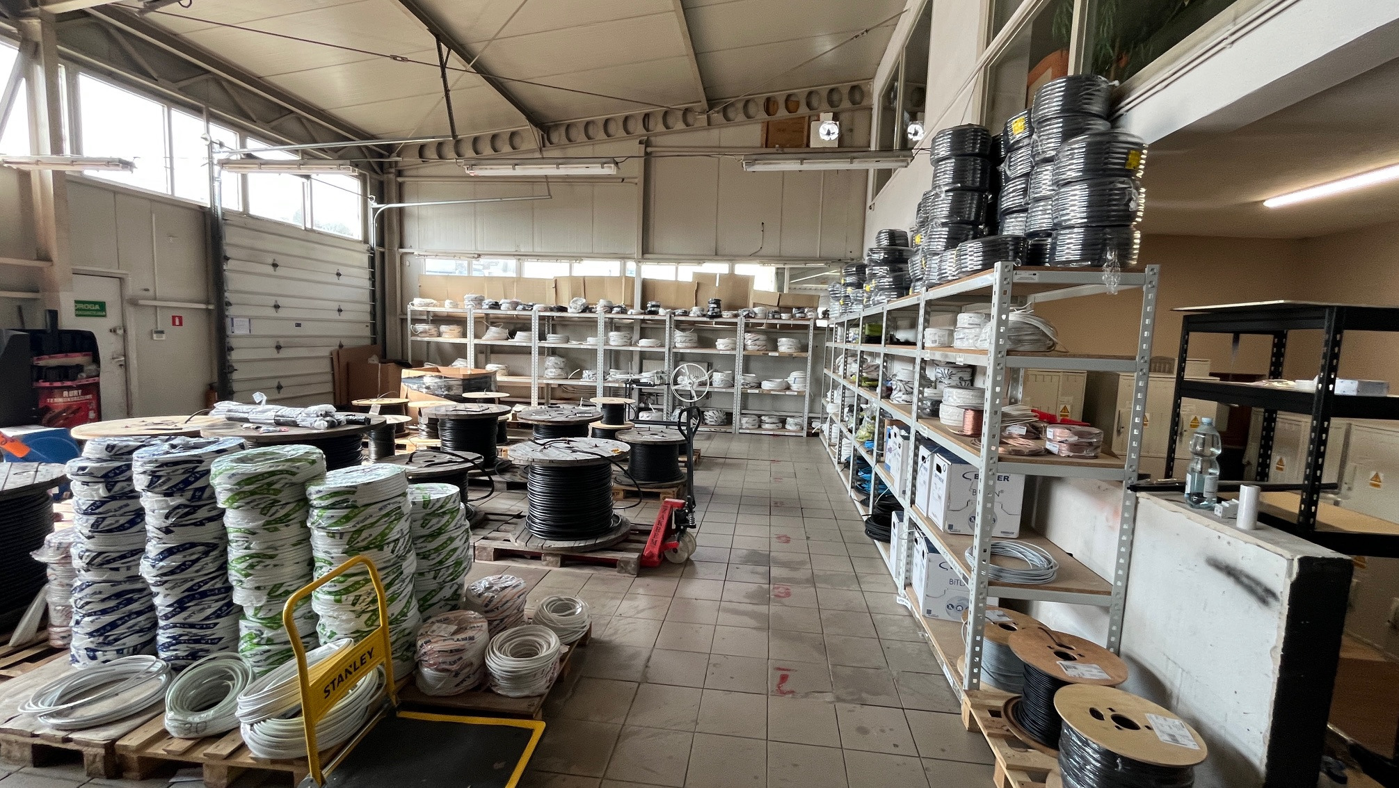 Wnętrze magazynu z kablami i przewodami na regałach i paletach. Widoczne rolki kabli w różnych kolorach i rozmiarach. Oświetlenie magazynowe, metalowe regały.